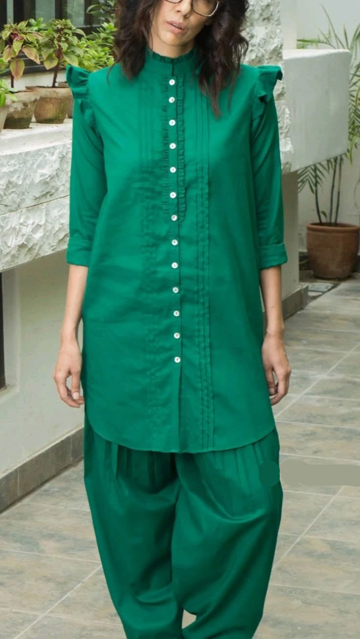 A woman wearing a green kurta and matching wide-leg pants set, standing in an outdoor patio setting with potted plants and tiled flooring.